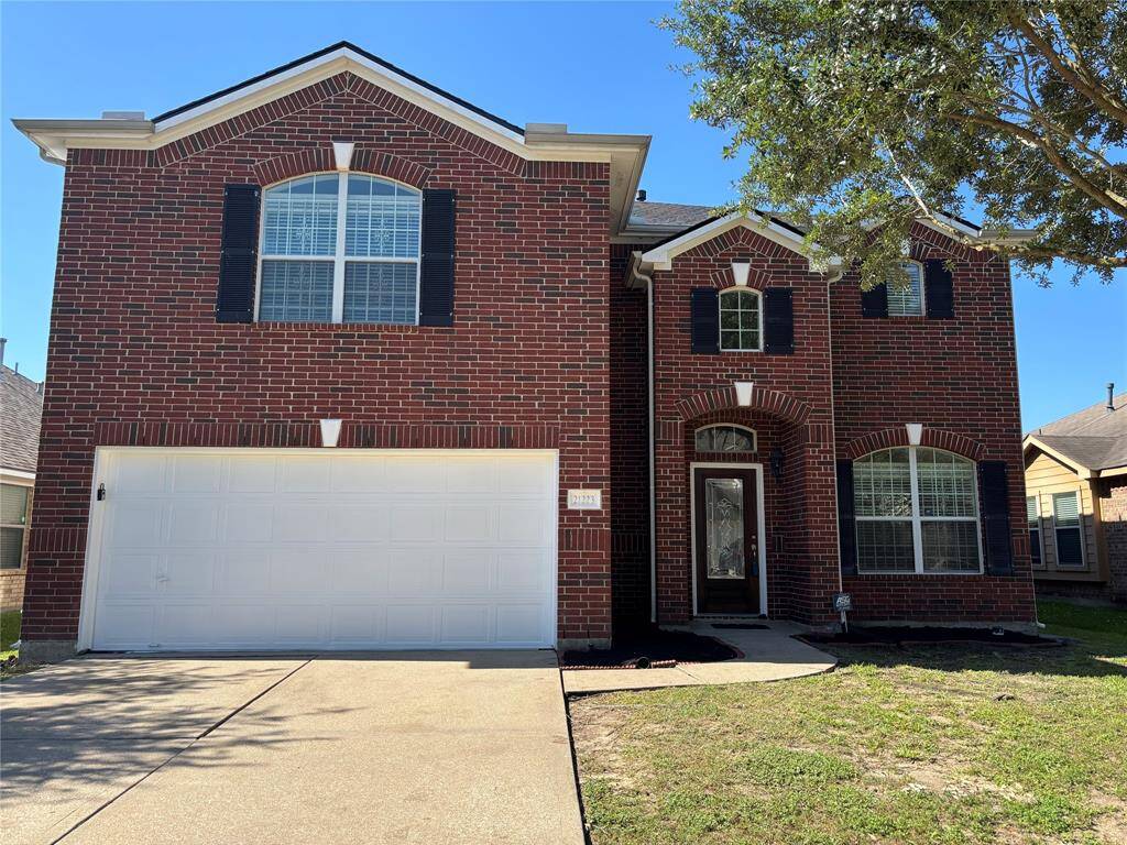 This is a two-story brick home featuring a large arched window above the garage and black shutters. It has a spacious driveway leading to a double garage and an inviting front entrance with decorative glass. The yard is partially shaded by a tree.