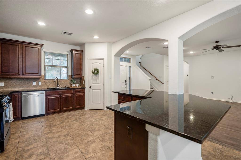 This photo showcases a spacious open-concept kitchen with dark wood cabinets, granite countertops, and stainless steel appliances. The room flows into a living area with a ceiling fan and visible stairway, featuring tile and wood flooring for a modern feel.