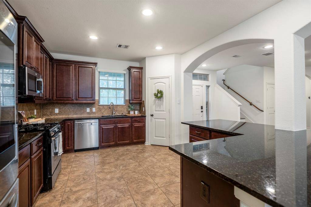 Spacious kitchen with dark wood cabinets, granite countertops, and stainless steel appliances. Features an open layout with tile flooring and an archway leading to the staircase. Ideal for modern living and entertaining.