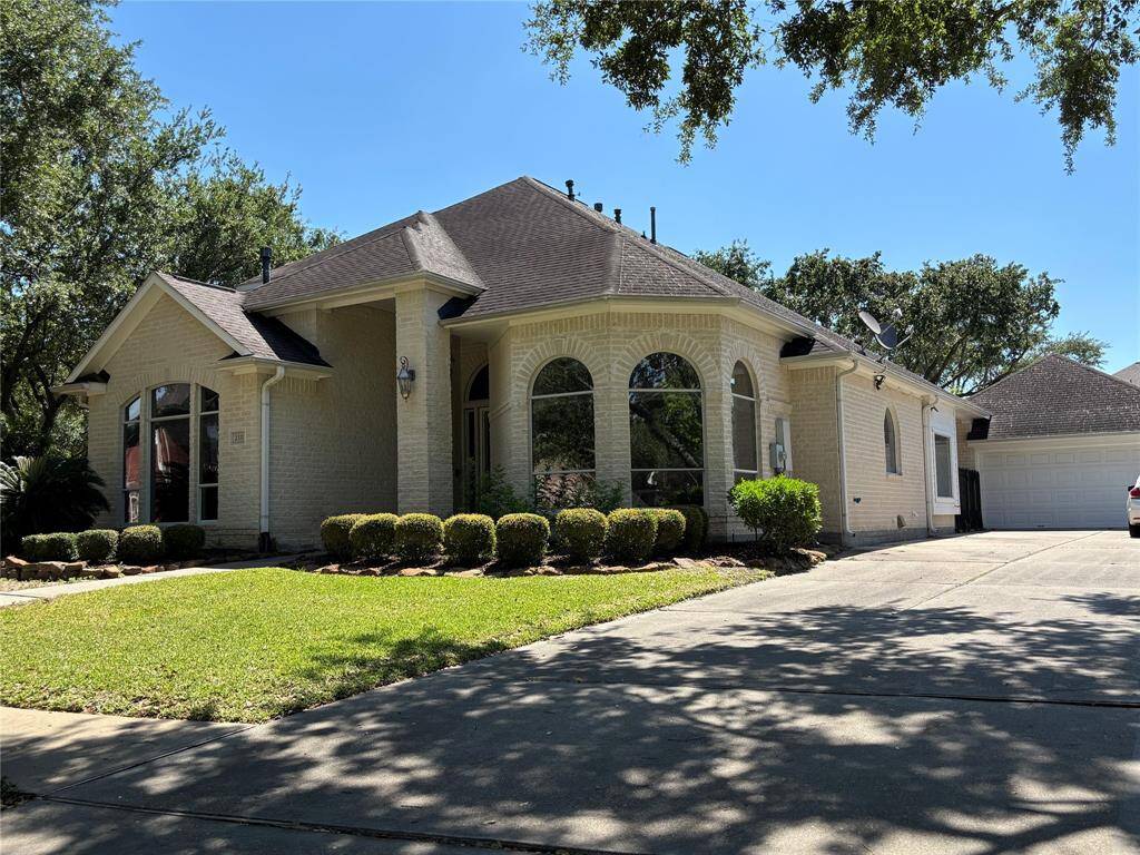 This charming single-story home features a light brick exterior, large arched windows, and a spacious driveway leading to a double garage. The well-maintained front yard is enhanced with manicured shrubs and mature trees, providing a welcoming curb appeal.