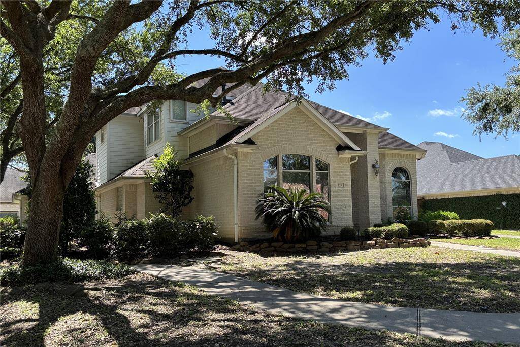 Charming two-story home with a light brick exterior, large arched windows, and a well-maintained front yard featuring mature trees and shrubs. The property offers a welcoming and shaded entrance.