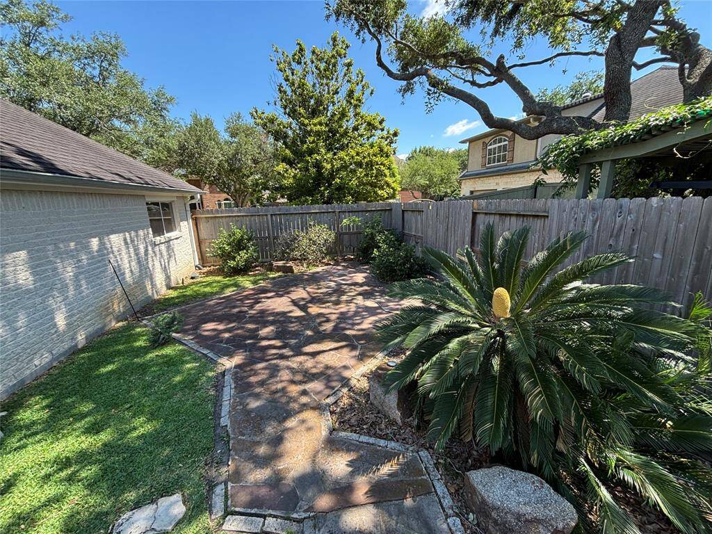 This photo showcases a backyard with a mix of paved and grassy areas, surrounded by a wooden fence. It features mature trees providing shade, a lush fern-like plant, and well-maintained landscaping, offering a serene outdoor space for relaxation.