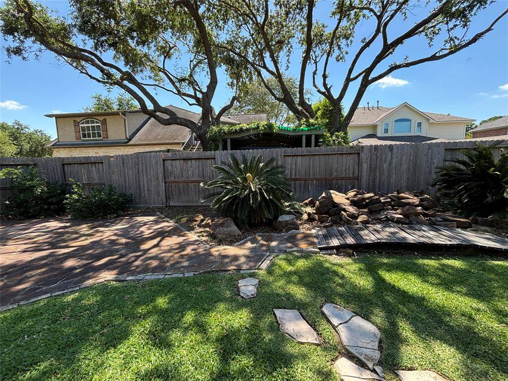 A well-maintained backyard with a stone patio, lush greenery, and a privacy fence. The house features a brick exterior and a detached garage, offering a mix of open space and shade from surrounding trees.