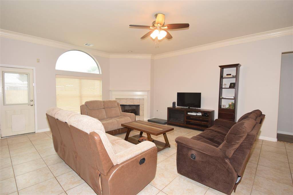 Spacious family room featuring double crown molding, tile floors, corner fireplace, ceiling fan with light fixture, door to the patio area and a breakfast bar opening to the kitchen.
