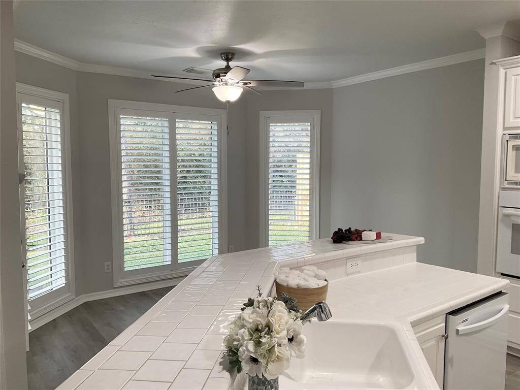 Bright and inviting kitchen area with large windows featuring plantation shutters, offering plenty of natural light. It includes a white-tiled countertop with a built-in sink, new dishwasher and garbage disposal. Elegant crown molding and trim enhance the custom home feelings.