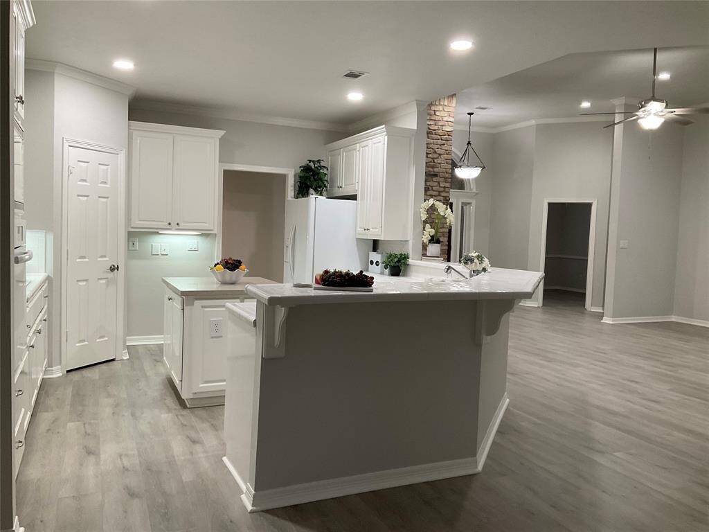 This photo showcases a modern, open-concept kitchen and living area. The kitchen features sleek white cabinetry, a large island with a sink, and stainless steel appliances. The space is well-lit with recessed lighting and has light wood flooring, creating a bright and inviting atmosphere.  You will love the breakfast bar area for additional seating.