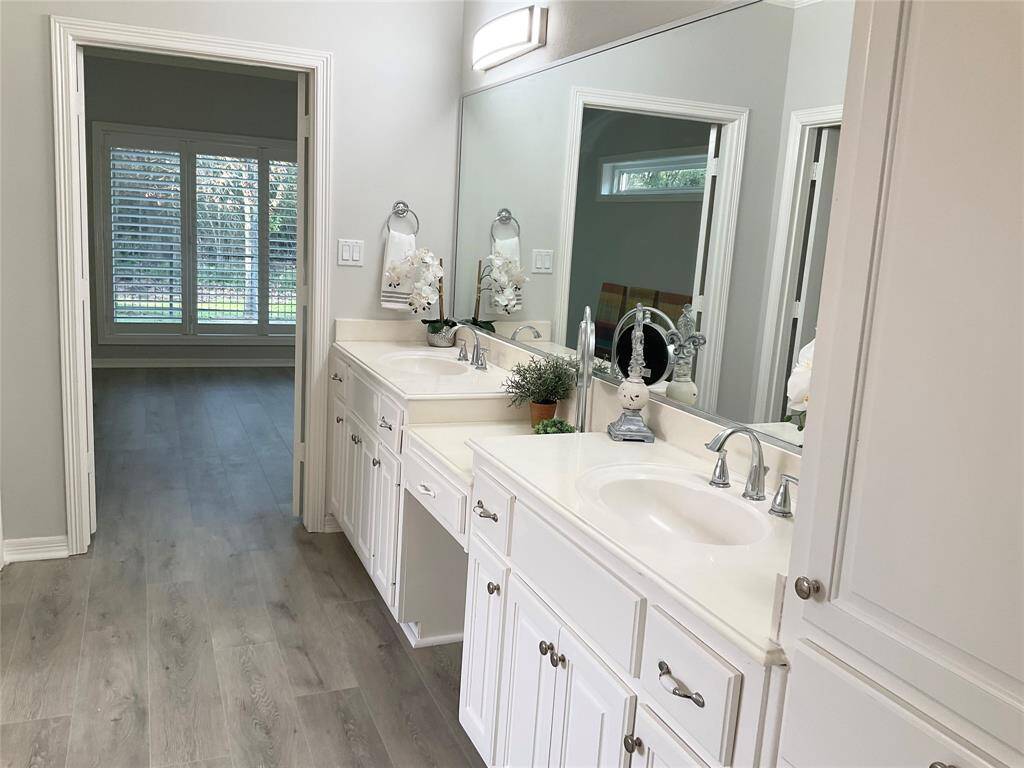 This bathroom features dual sinks with a large mirror, white cabinetry, and light wood flooring. It connects to a room with large windows and plantation shutters, offering ample natural light.