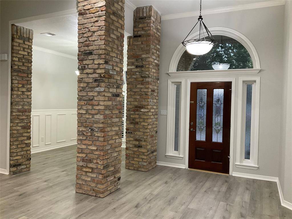 This entryway features a rich wooden front door with decorative glass panels, framed by brick columns and large windows. The space has high ceilings, modern light fixtures, and light wood flooring, creating a welcoming and elegant atmosphere.
