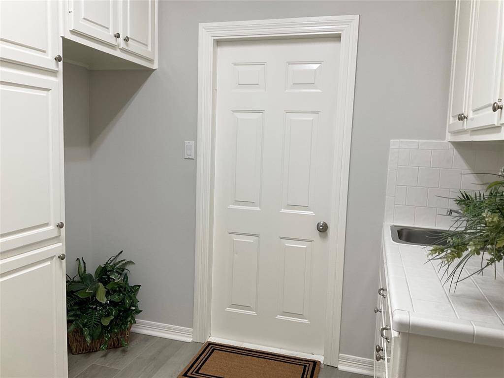 Bright utility room with white cabinetry, a sink, and new flooring. Lots of cabinetry in the utility room with a sink and room for another refrigerator in the space on the left.  The door leads to a spotless, oversized garage with epoxy paint flooring.  The home or streets have never flooded in this community.
