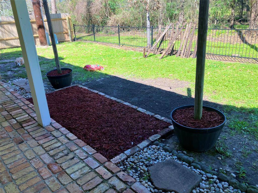 This photo shows a backyard with a brick patio and a landscaped area featuring mulch and potted plants.   A great spot for a fire pit and roasting marshmallows. There's a grassy yard beyond the patio enclosed by a fence, offering privacy and space for outdoor activities.  The private 5. acres behind the home is owned by MUD 19 and will not be developed.