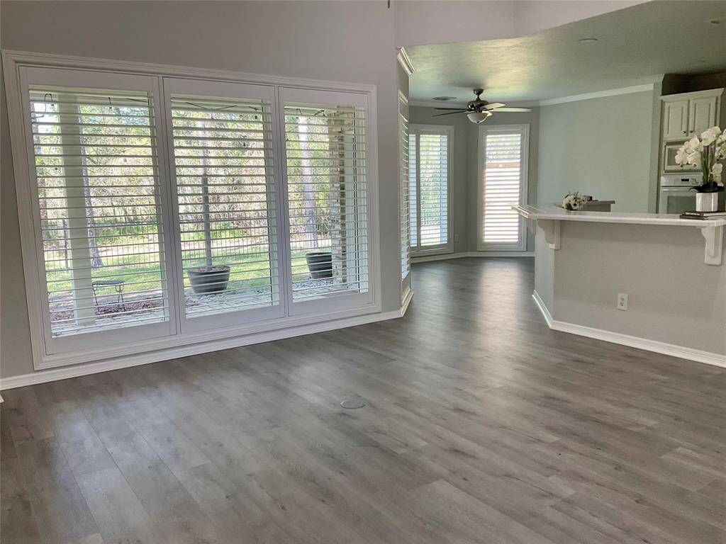This spacious, light-filled room features large windows with plantation shutters, modern, new flooring, and a view of the private outdoors. The open layout flows into a cozy dining area with a ceiling fan, adjacent to a kitchen with a breakfast bar that will allow for at least 6 bar stools.