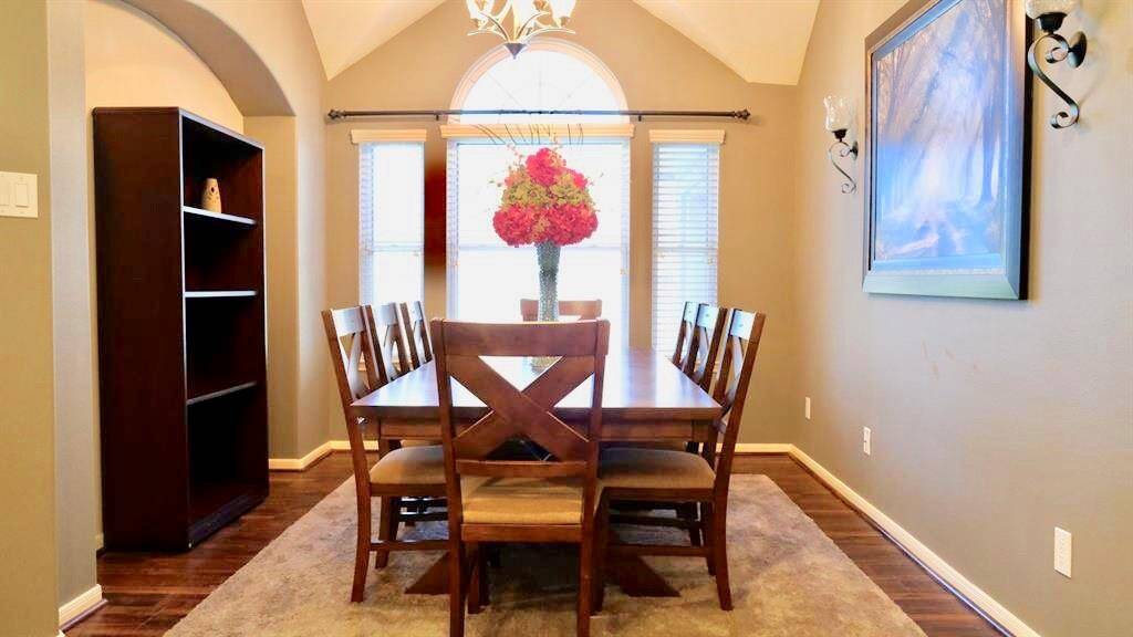 Nice dining room with warm lighting and a vaulted ceiling. It features a wooden dining table with six chairs, a large window with natural light, a decorative centerpiece, and wall sconces. The room has neutral walls, a soft area rug, and hardwood flooring, creating an inviting atmosphere.