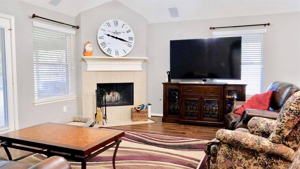Cozy living room with a vaulted ceiling, featuring a fireplace with a large decorative clock above it. The space includes a comfortable seating area with a TV on a wooden cabinet, hardwood floors, and plenty of natural light from two windows.