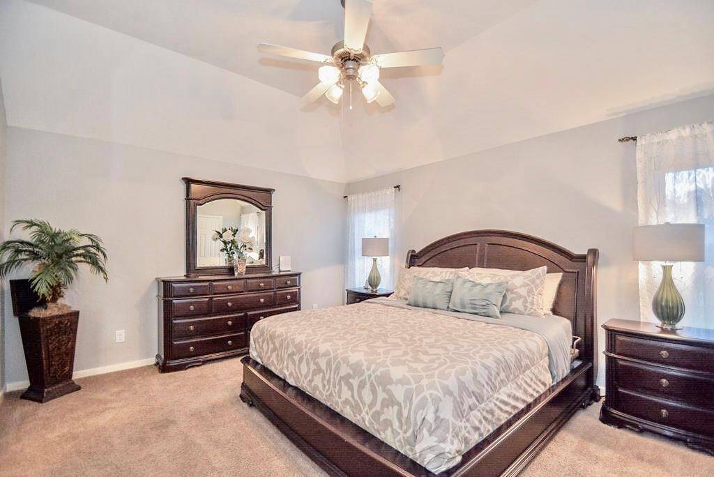 This spacious bedroom features a vaulted ceiling with a ceiling fan, a large bed with elegant wooden furniture, including matching dressers and nightstands. Soft lighting and neutral colors create a warm, inviting atmosphere, perfect for relaxation.