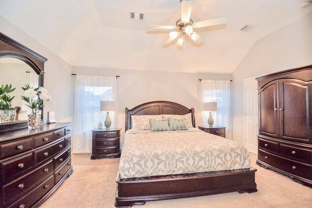 Bright and spacious bedroom with vaulted ceilings, featuring elegant dark wood furniture, including a dresser, nightstands, and an armoire. The room is well-lit with natural light from two windows, complemented by a ceiling fan and soft, neutral decor.