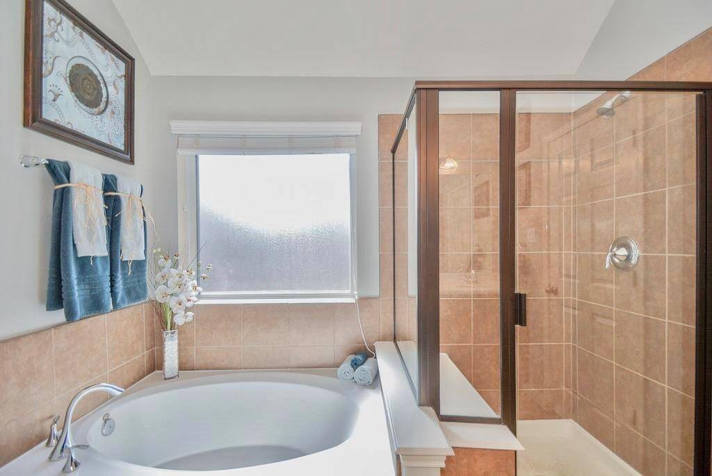 This bathroom features a sleek glass-enclosed shower with beige tiles, a large soaking tub, and a frosted window that brings in natural light. The decor includes blue towels, floral accents, and a framed artwork for a touch of elegance.