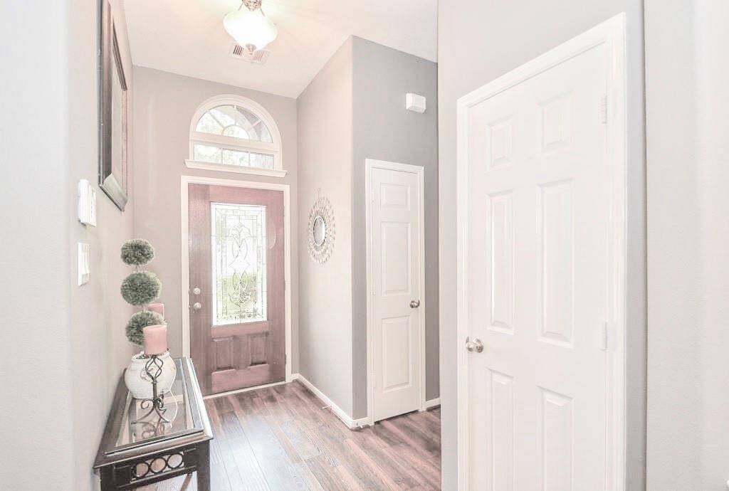 Bright and welcoming entryway with a decorative glass front door, high ceiling, and elegant chandelier. It features light gray walls, hardwood flooring, and a console table with decor accents, creating a modern and inviting atmosphere.