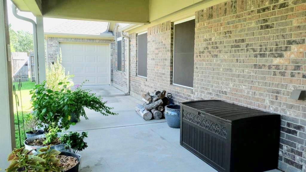 Covered patio area with a brick exterior, featuring potted plants and a storage bench. It leads to a garage and has a small stack of firewood, perfect for outdoor relaxation and storage.