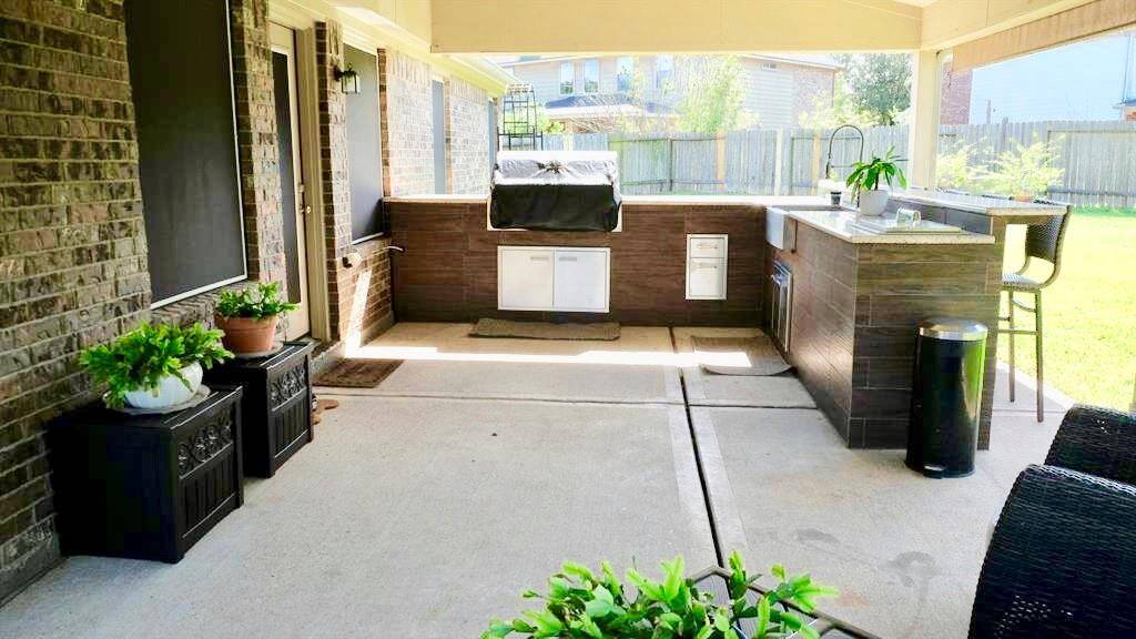 Covered outdoor patio with a built-in kitchen, including a grill and sink. It features a brick design, ample counter space, and seating, overlooking a fenced backyard—ideal for entertaining.