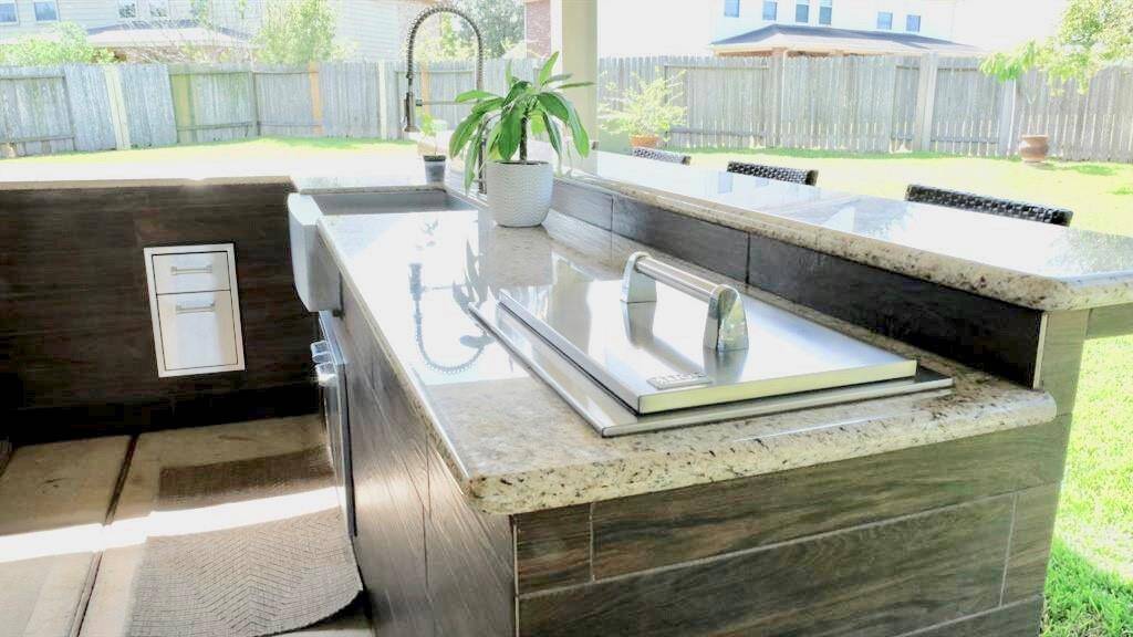 Modern outdoor kitchen with granite countertops, a built-in grill, and a sink. It's part of a spacious patio area, ideal for entertaining, with a view of a fenced backyard.