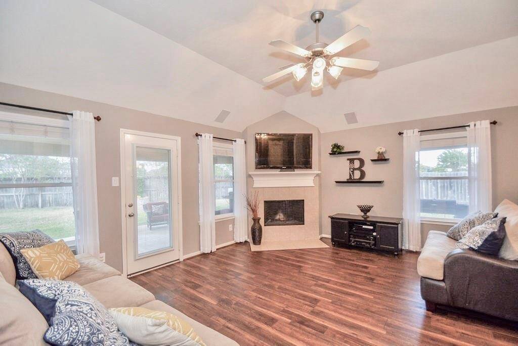 Spacious living room features high ceilings with a ceiling fan, large windows for natural light, a cozy fireplace, and hardwood floors. The open layout includes a glass door leading to the backyard, creating a bright and inviting atmosphere.