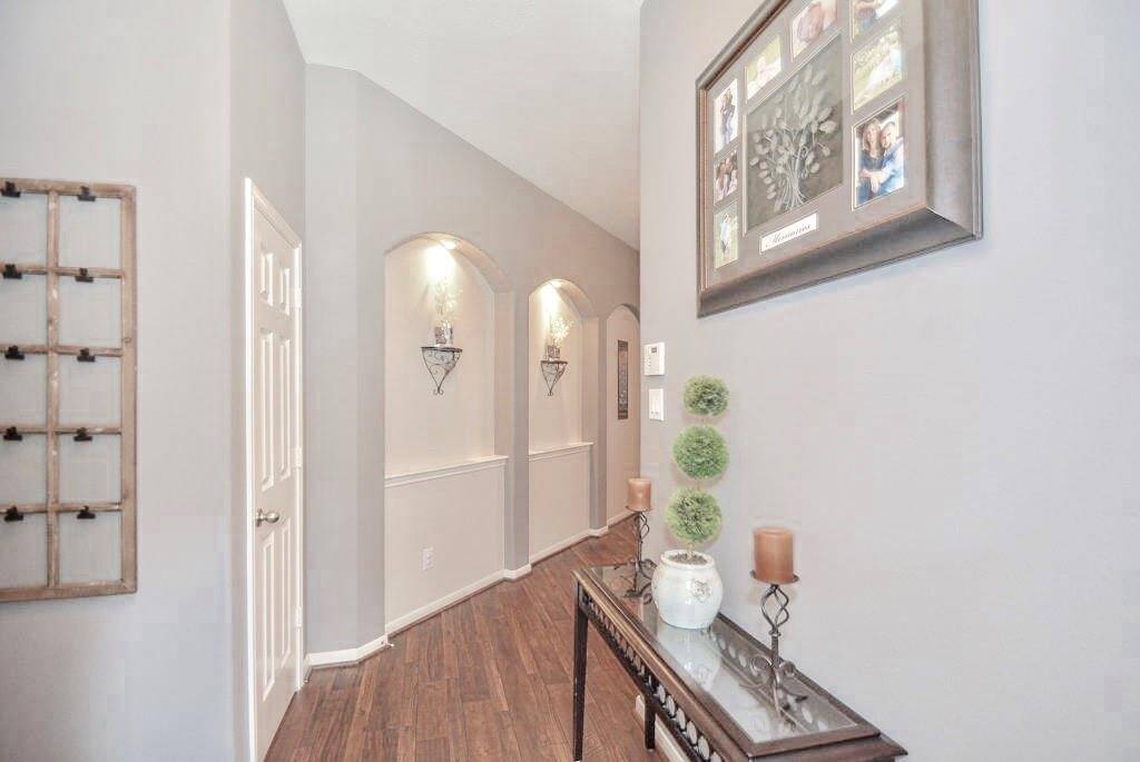 This hallway features elegant hardwood flooring, soft gray walls, and decorative niches with lighting. A console table with greenery and candles adds charm, while framed art enhances the aesthetic appeal.