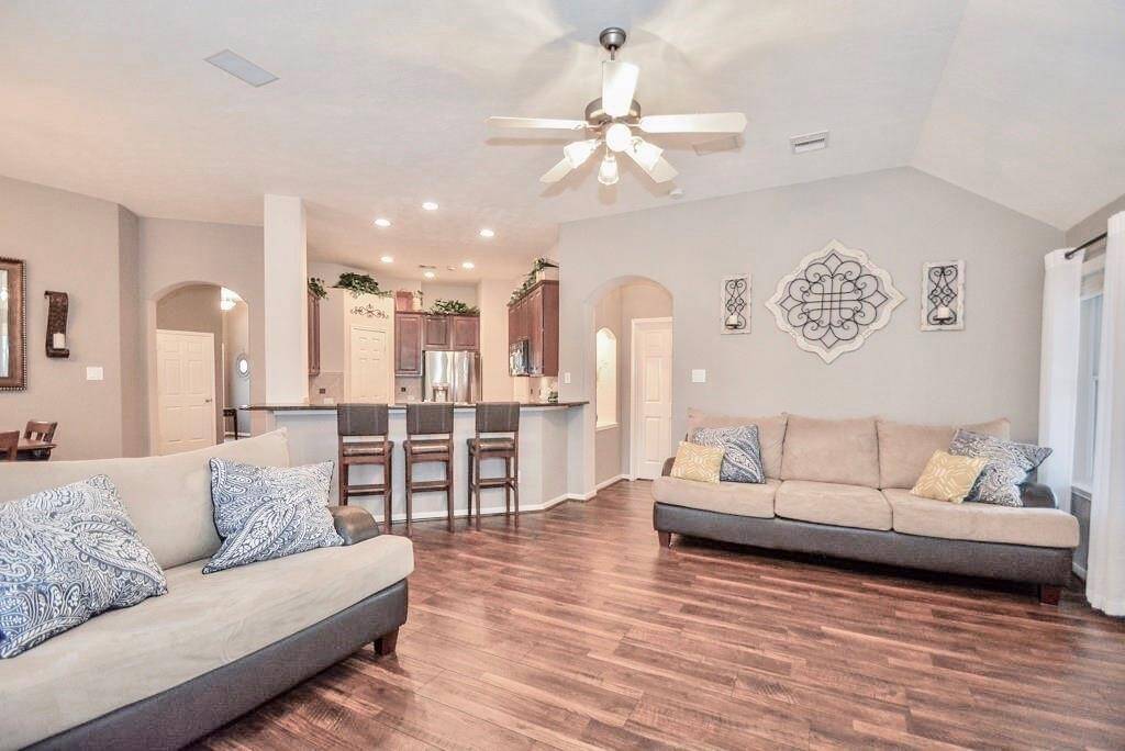 Spacious living area features elegant hardwood floors, neutral walls, and a ceiling fan. The open layout seamlessly connects to a modern kitchen with a breakfast bar and stainless steel appliances. Cozy seating and decorative accents provide a welcoming atmosphere.