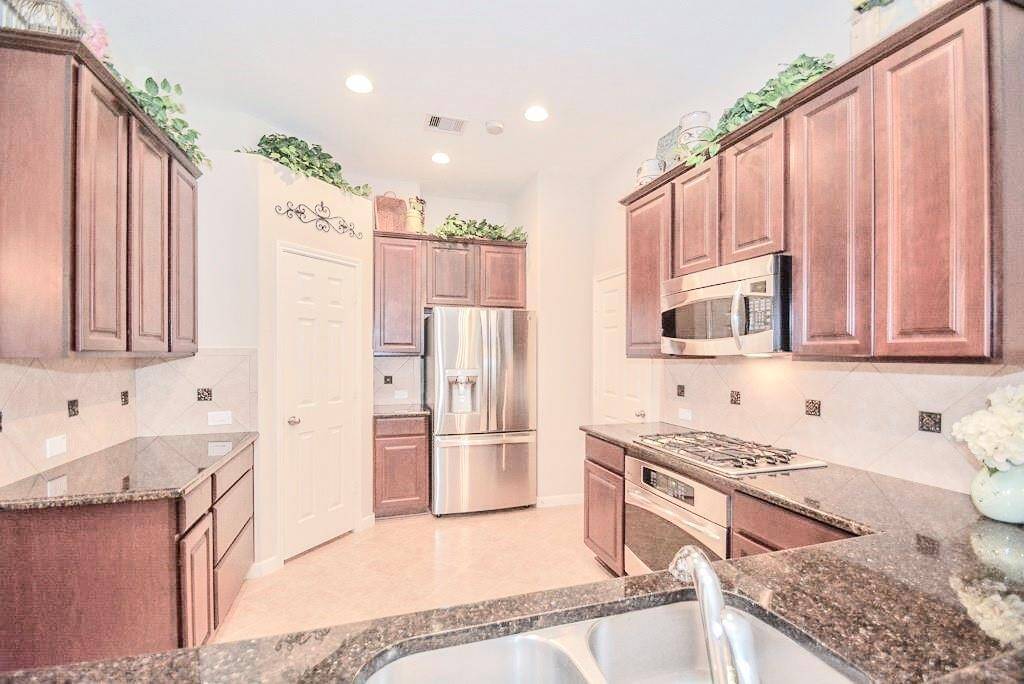 The Kitchen features warm wooden cabinetry, granite countertops, and stainless steel appliances, including a built-in microwave and a double-door fridge. The space is accented with decorative tile backsplash and recessed lighting, creating a bright and inviting atmosphere.