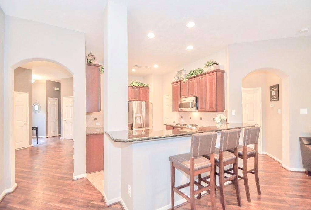 Modern kitchen with granite countertops, wooden cabinets, and a breakfast bar with seating for three. The space is well-lit with recessed lighting and features arched doorways, leading into adjacent rooms. The flooring is a rich, dark wood, adding warmth to the area.
