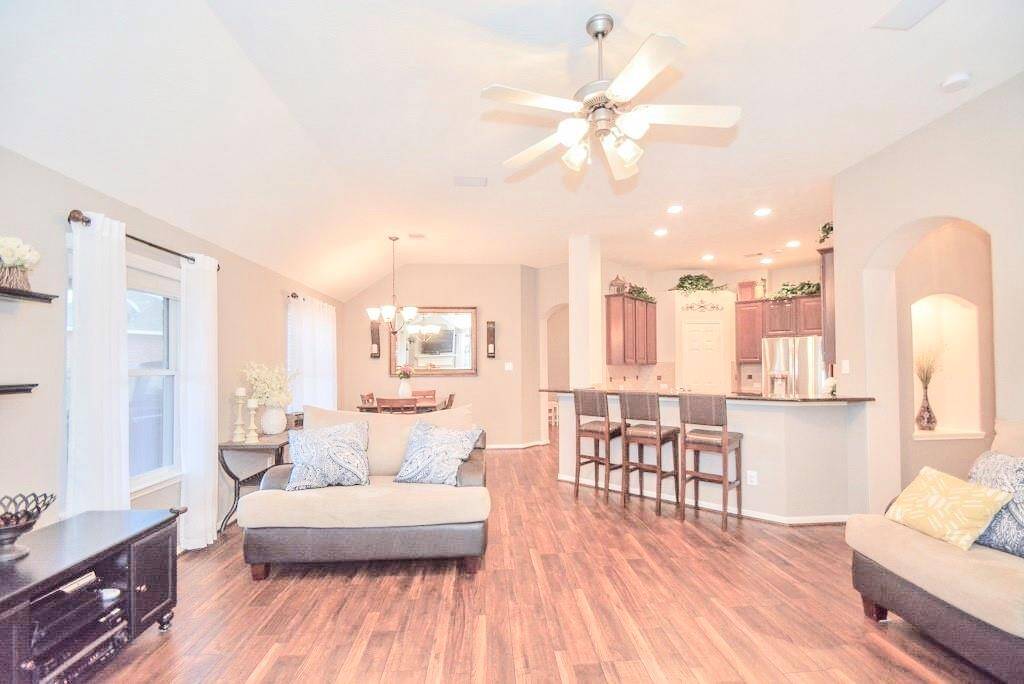 Spacious open-plan living area with hardwood floors, modern ceiling fan, and abundant natural light. The space seamlessly connects to a kitchen with a breakfast bar and seating, featuring elegant cabinetry and stainless steel appliances. Cozy seating and tasteful decor complete the inviting ambiance.