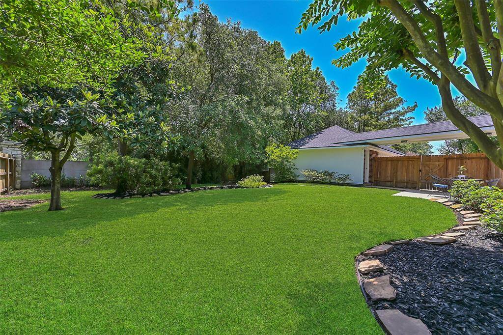Lush backyard oasis with mature trees, manicured landscaping, and privacy provided by a tall rear fence lined with trees. The space is ideal for play, pets, or relaxing under the shade, with direct access to the garage and patio seating area.
