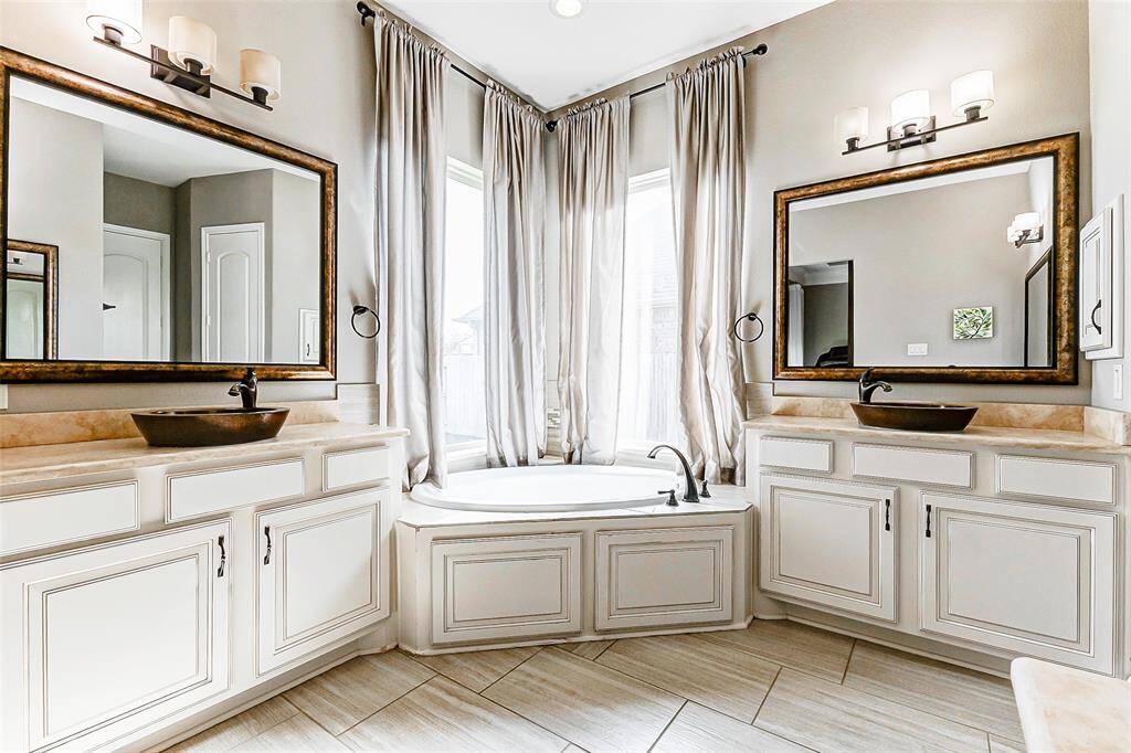The ensuite features multiple vanities and storage options, two of which are equipped with elegant vessel sinks, framed mirrors, and upgraded lighting fixtures. The soaking tub, perfectly positioned between the vanities, is bathed in natural light from two large windows that frame the serene view, enhancing the spa-like ambiance.