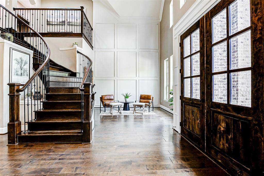 A striking wooden staircase with elegant iron balusters serves as a stunning focal point, perfectly complemented by grand double entry doors that elevate the home's sense of arrival. Just beyond, the formal living room features custom wall paneling, adding depth and sophistication to the space.