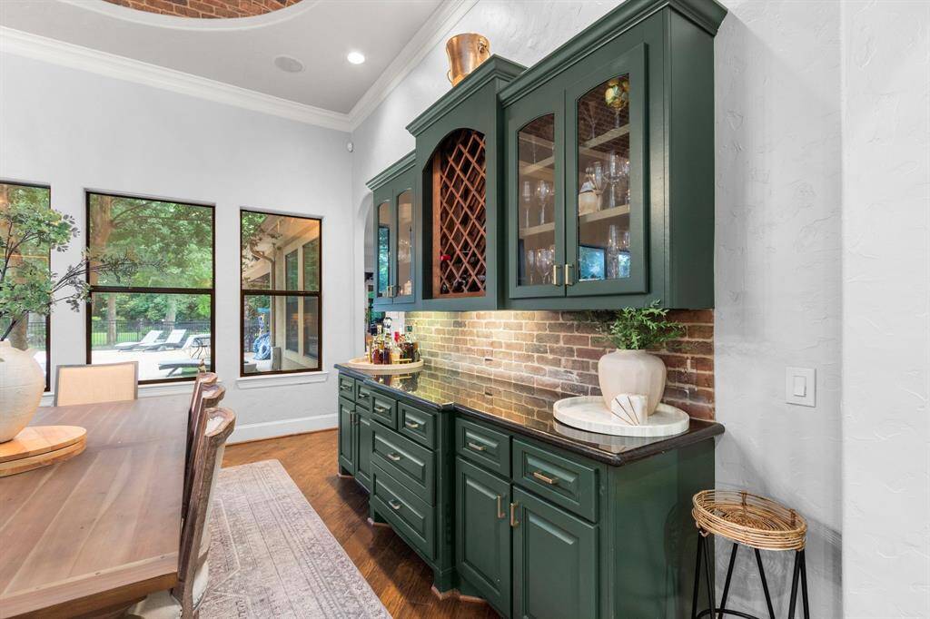 This inviting dining area features rich green cabinetry with a built-in wine rack and glass display. A brick backsplash adds rustic charm, while large windows offer backyard views.