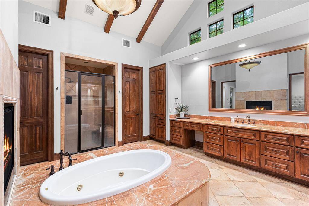 The primary bathroom features a luxurious soaking tub, large vanity with ample storage, and a separate shower. It boasts high ceilings with exposed beams, a cozy fireplace, and elegant wood finishes, creating a warm and inviting atmosphere.
