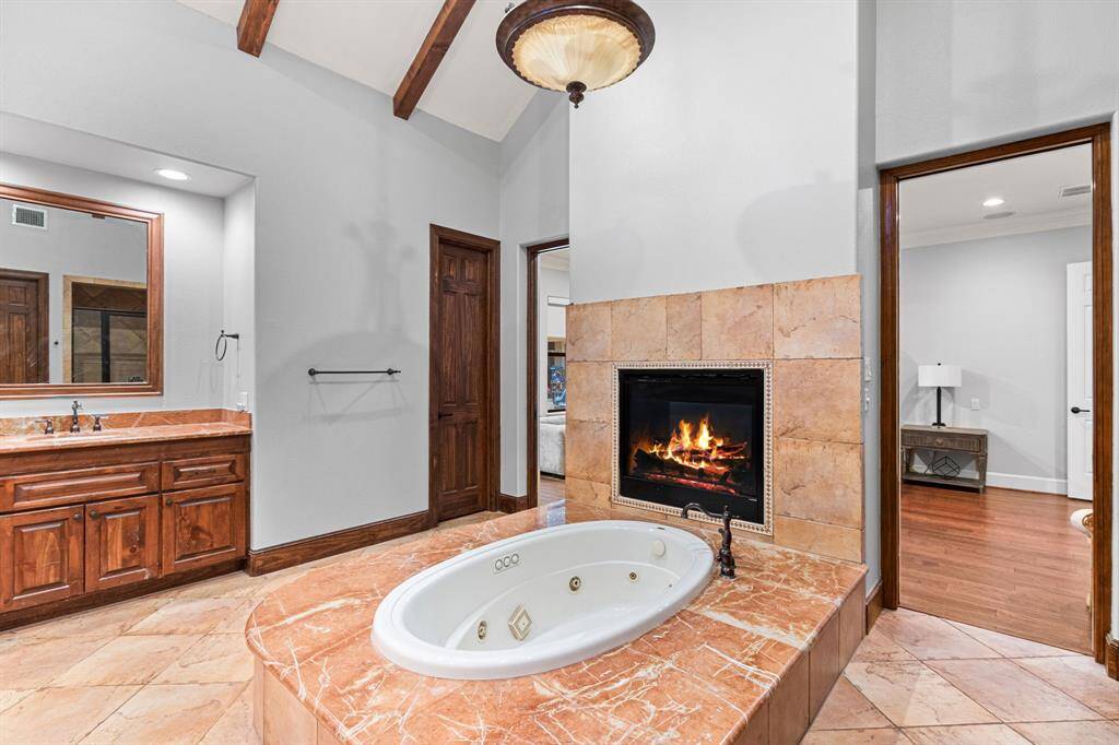 This luxurious bathroom features a sunken jetted tub with a warm, tile surround and an elegant double-sided fireplace. The space is accented with wooden cabinets and beams, providing a cozy and inviting atmosphere.