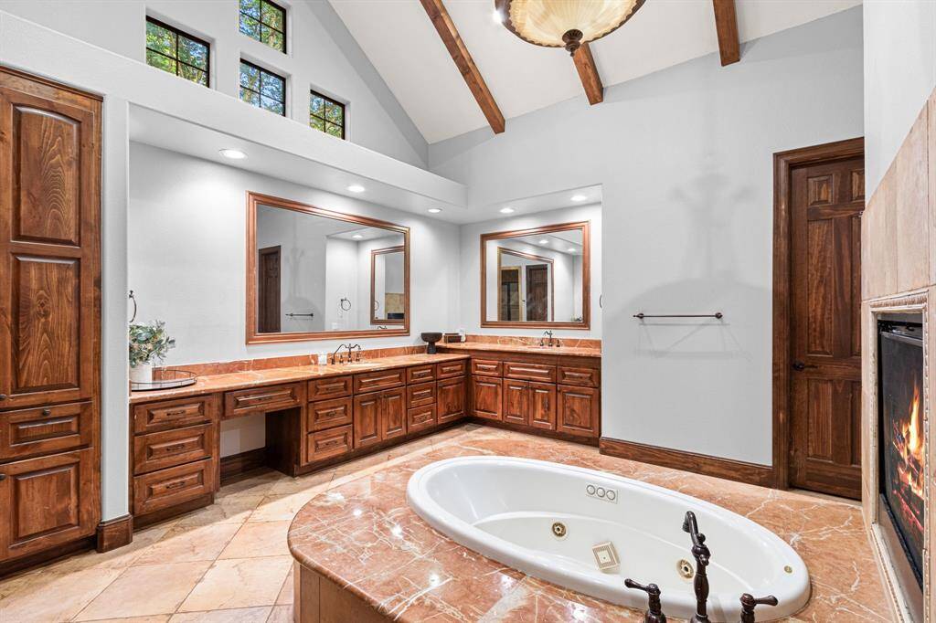This luxurious bathroom features a high vaulted ceiling with exposed wooden beams, elegant wooden cabinetry, and a large soaking tub set in marble. It includes spacious dual vanities, a cozy fireplace, creating a serene and sophisticated retreat.