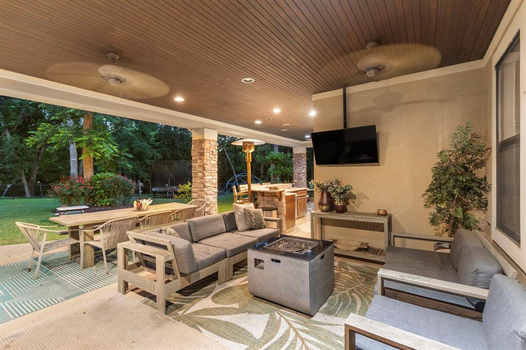 This photo showcases a spacious and inviting outdoor patio, featuring comfortable seating around a modern fire pit, a wall-mounted TV, and a dining area. The ceiling fans and recessed lighting enhance the ambiance, while the view of a lush, landscaped backyard provides a serene backdrop. Perfect for entertaining or relaxing.