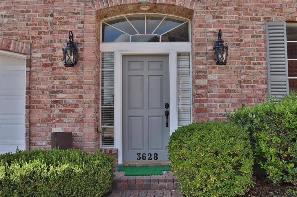 Charming brick home entry with a classic gray door, flanked by elegant lanterns and sidelights. Features a welcoming arch transom window and well-maintained shrubs, enhancing curb appeal.