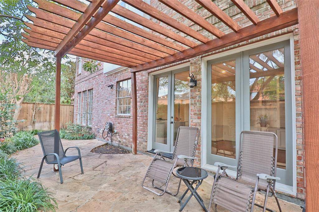 This inviting patio features a charming wooden pergola and a stone floor, perfect for outdoor relaxation. Surrounded by greenery, it includes comfortable seating and provides easy access to the home through large glass doors, enhancing the indoor-outdoor living experience.
