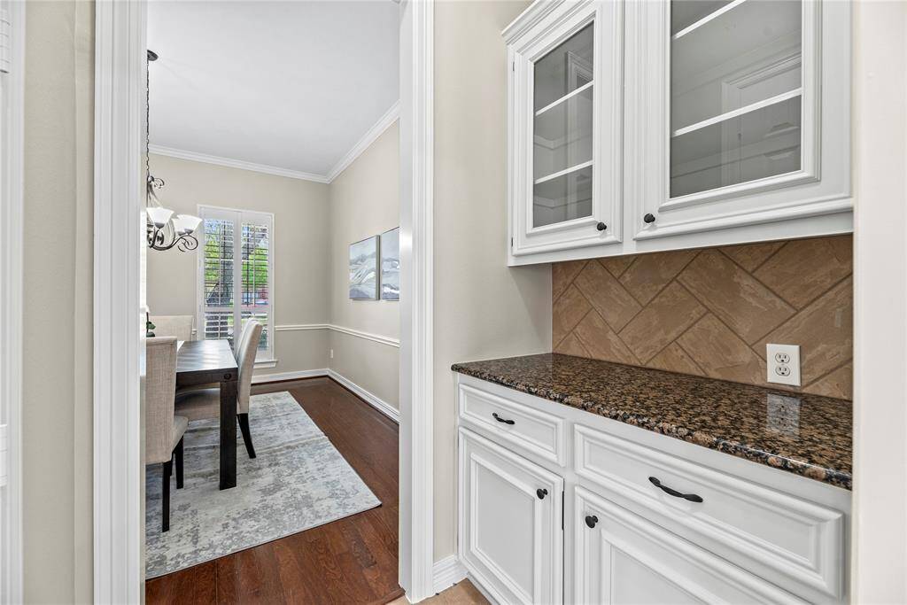 The butler's pantry featuring granite countertops, white cabinetry with glass doors, and a stylish tile backsplash.