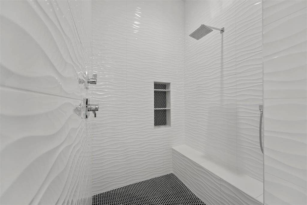 The oversized walk-in shower stuns with wave- textured tile, custom bench, rainfall shower head, and seamless glass enclosure