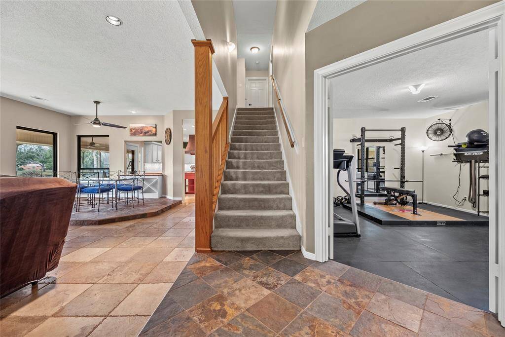 This home features a spacious open floor plan with a tiled entryway leading to a carpeted staircase. To the right, there's a well-equipped home gym. The living area is bright with large windows, and the adjacent dining space flows into a modern kitchen.