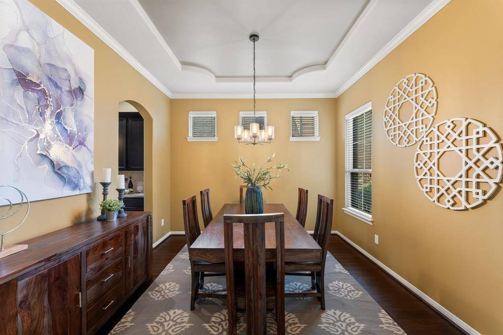 This elegant dining room showcases a combination of large and small windows for balanced natural light, detailed crown molding, a beautifully designed ceiling, rich wood flooring, and seamless access to the kitchen.