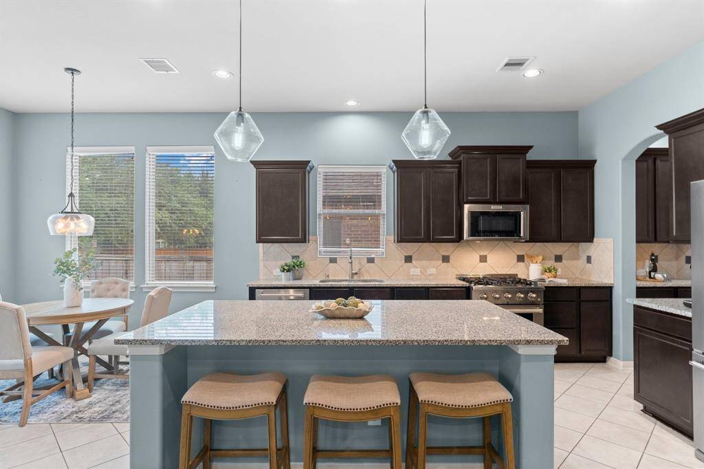 This open and spacious kitchen features a large granite island with additional seating, ample prep and cooking space, generous storage, sleek stainless steel appliances, recessed lighting, and stylish pendant lights.
