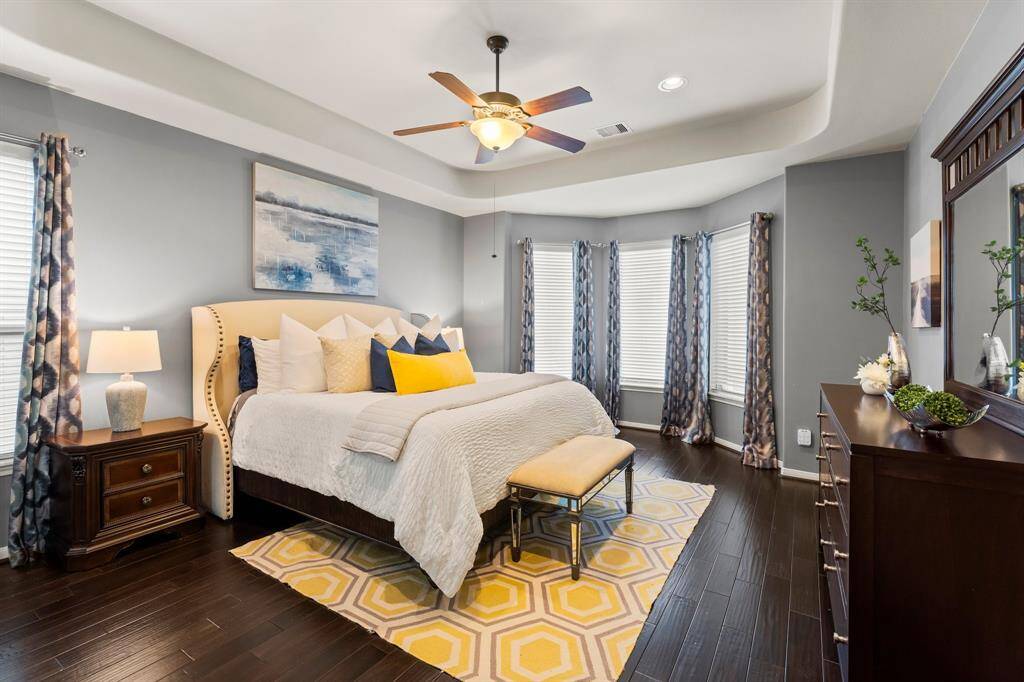 The beautiful and spacious primary bedroom features elegant tray ceilings, expansive windows that fill the room with natural light, beautiful wood flooring, and recessed lighting—creating a serene retreat perfect for relaxation.