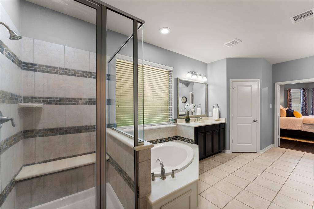The spacious primary bathroom features dual vanities, an oversized walk-in shower, and a separate jetted tub—all beautifully illuminated by a large frosted window, recessed lighting, and stylish vanity lights for a bright, spa-like ambiance.