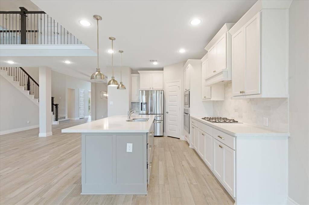 This photo showcases a modern kitchen with white cabinetry, stainless steel appliances, and a spacious island with pendant lighting. The open layout flows into a bright living area with a staircase, offering a contemporary and inviting atmosphere.