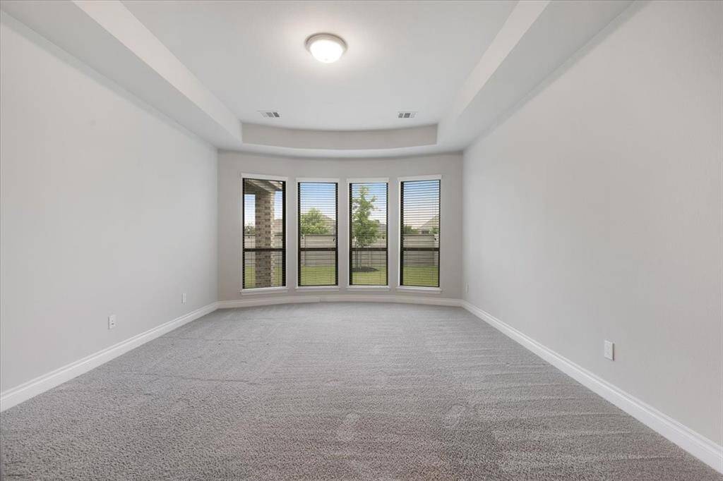 This room features a spacious design with light gray walls and carpet, a tray ceiling with recessed lighting, and a large bay window offering plenty of natural light and a view of the greenery outside.