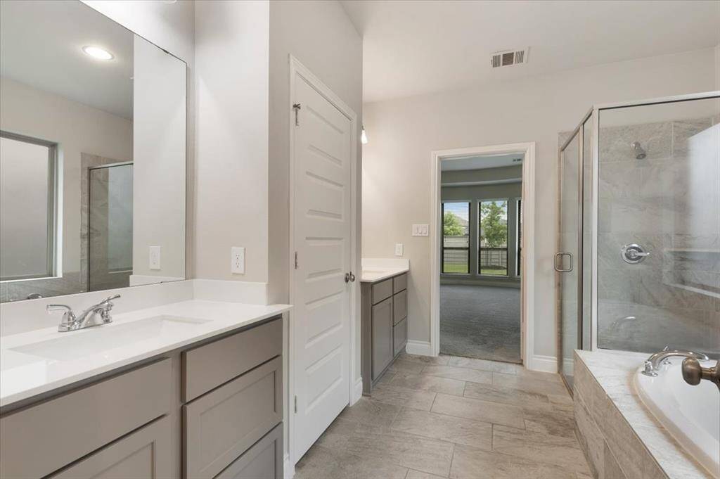 This modern bathroom features dual sinks with ample storage, a spacious glass-enclosed shower, and a large soaking tub. The neutral color palette and tile flooring create a clean, inviting atmosphere, with a view leading into a sunlit room with large windows.